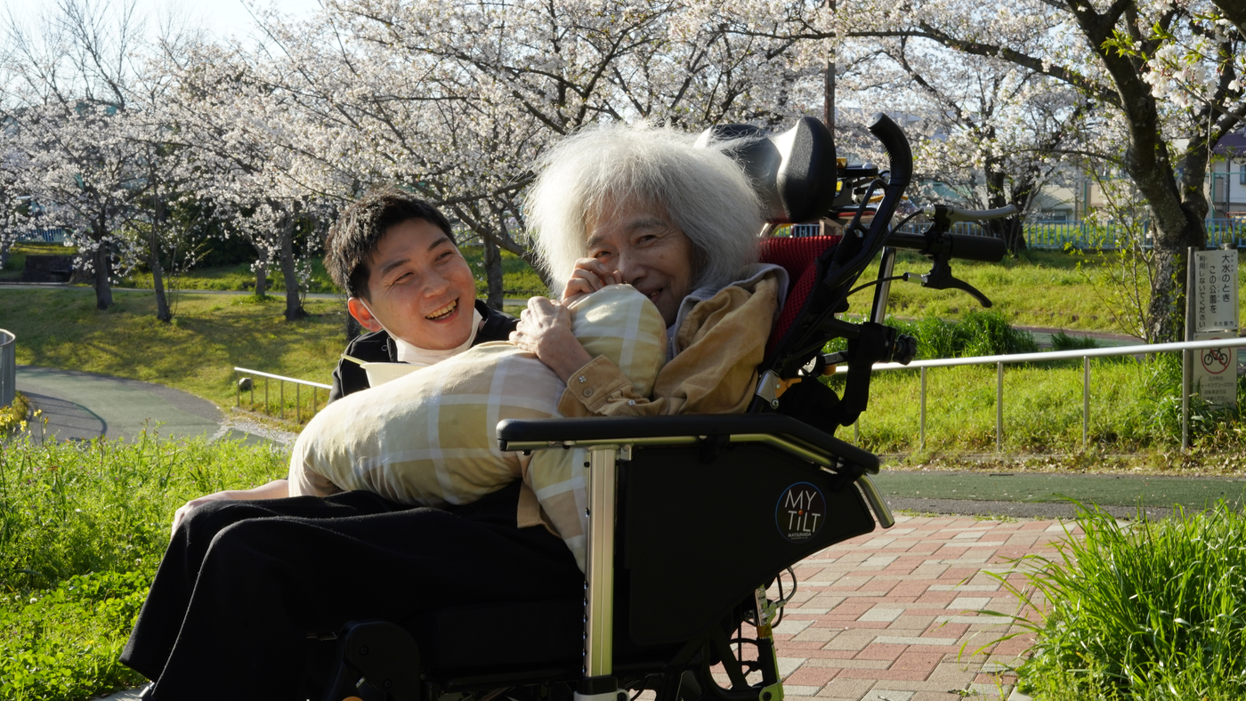 桜の下で車いすの入居者に笑顔で寄り添うナーシングホームかんなのスタッフ